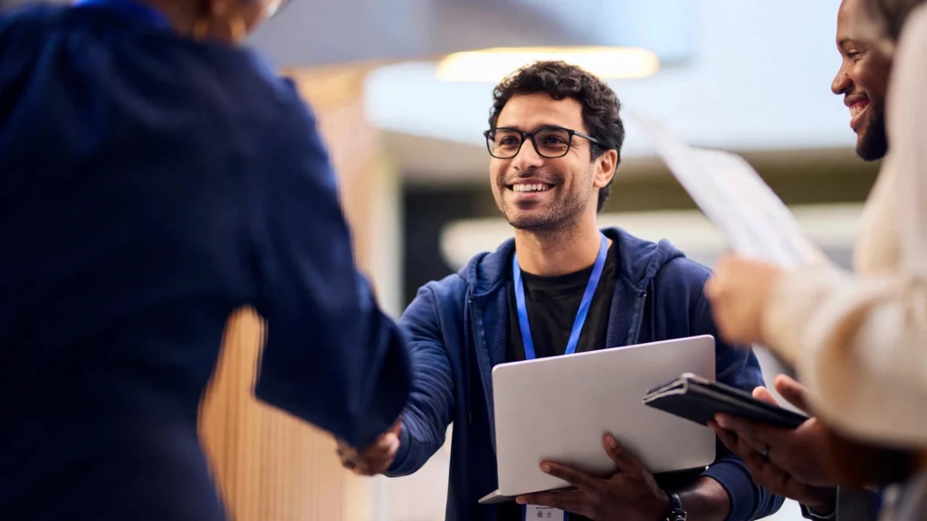 Lachende gedetacheerde IT-specialist met laptop schudt de hand van een nieuwe opdrachtgever via Morgan Black.