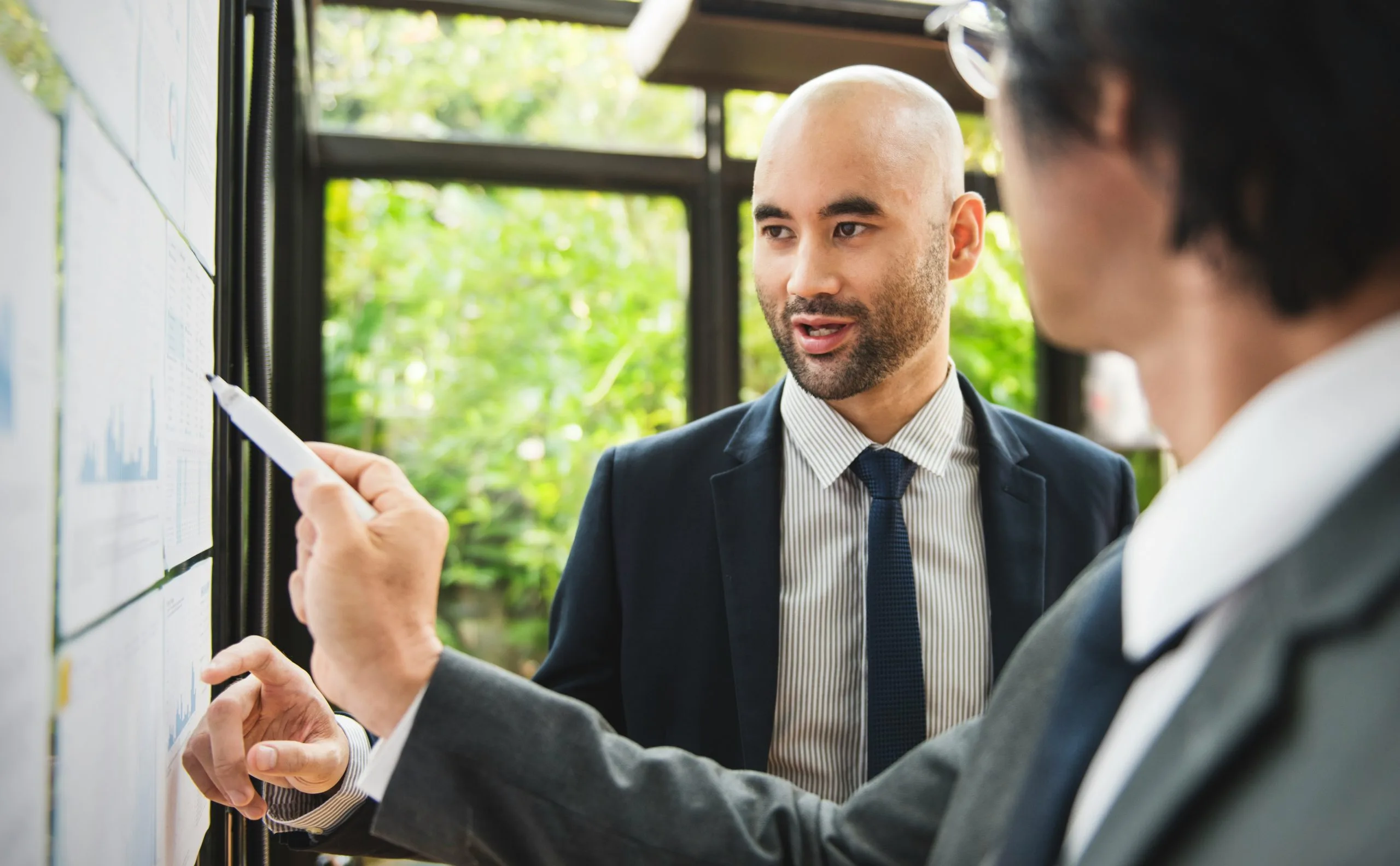 Twee data science en AI-professionals in een diepgaand technisch gesprek. Een wervingsspecialist wijst met een marker naar grafieken en gegevens op een kantoormuur, wat het nauwkeurige screeningproces voor IT- en AI-talentsourcing visualiseert.